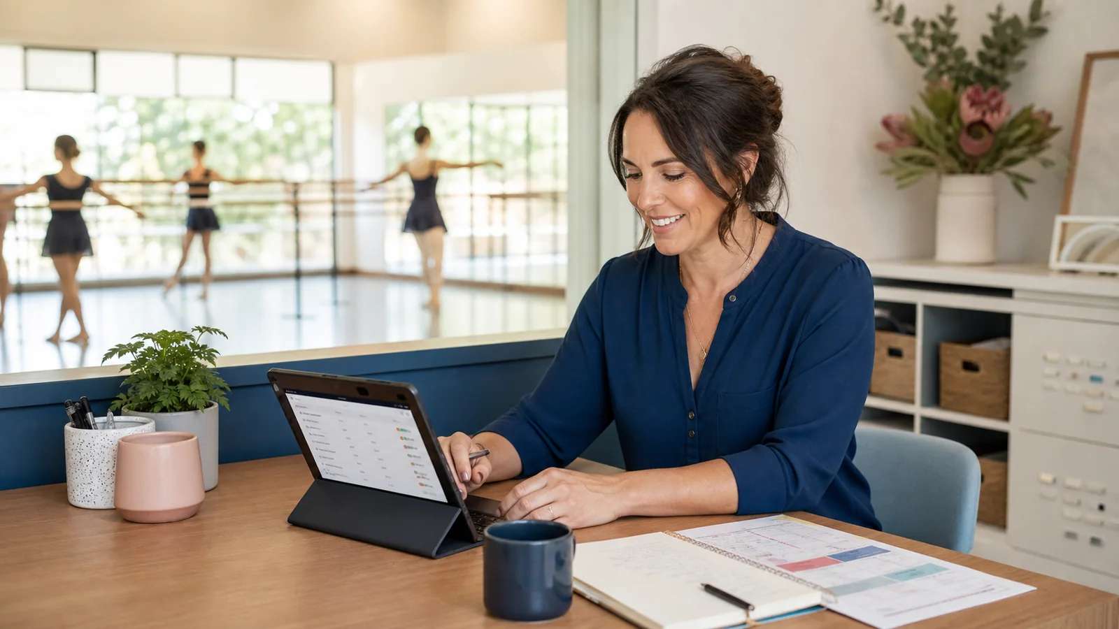 A studio owner reviewing the schedule on a tablet while a ballet class practises at the barre behind her